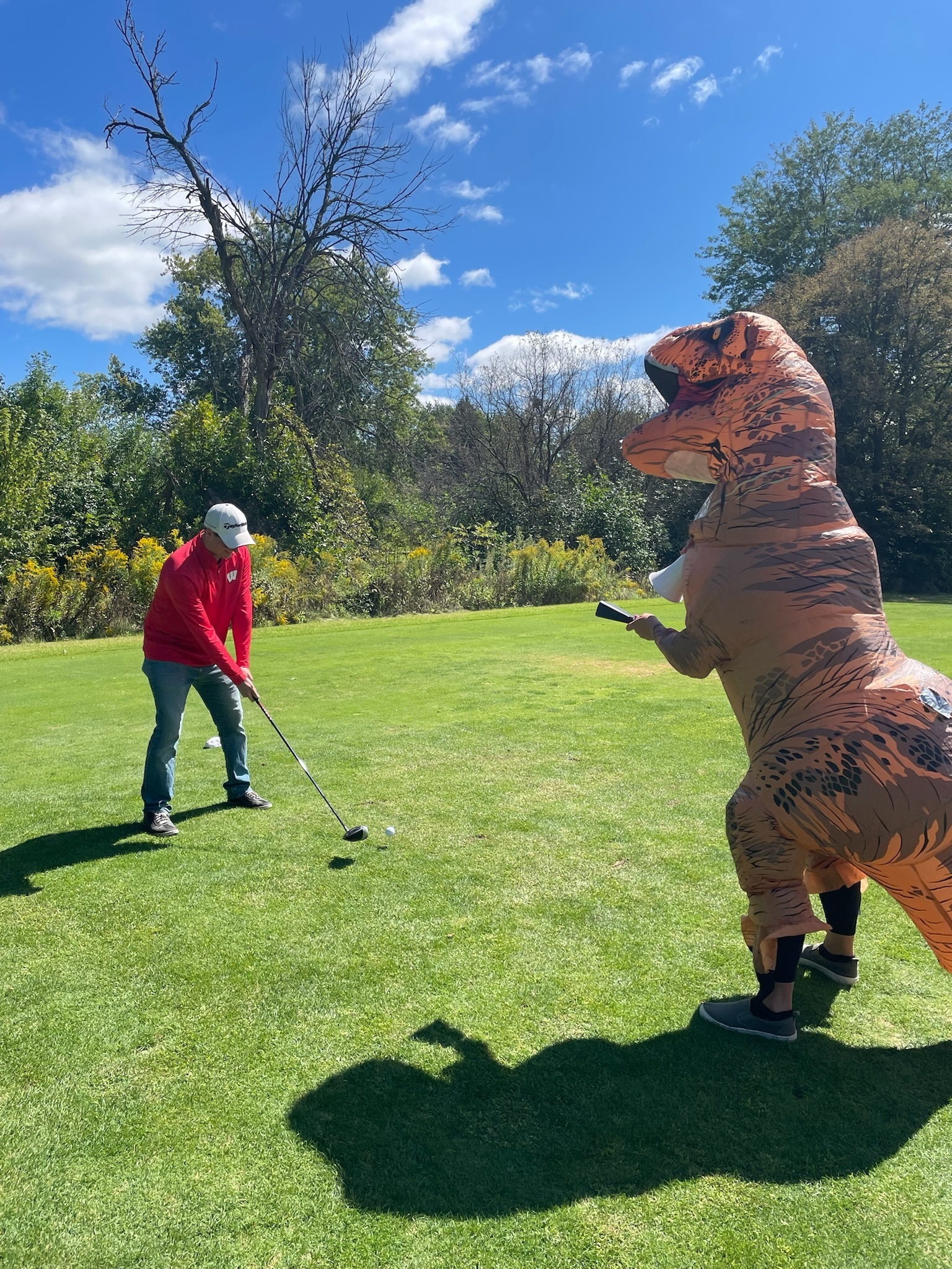 A golfer teeing off while someone in a dinosaur costume watches at Coop's Cup